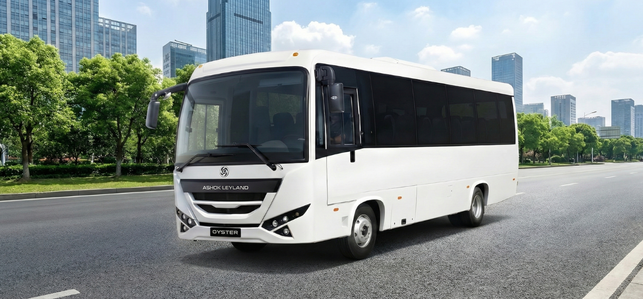 A white Ashok Leyland Oyster bus drives along a road with tall buildings