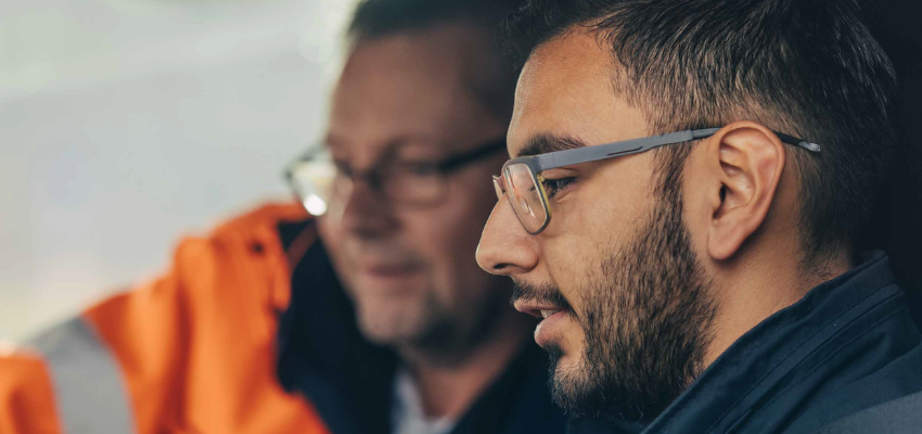 Two men in orange jackets and glasses are intently looking at a tablet together.