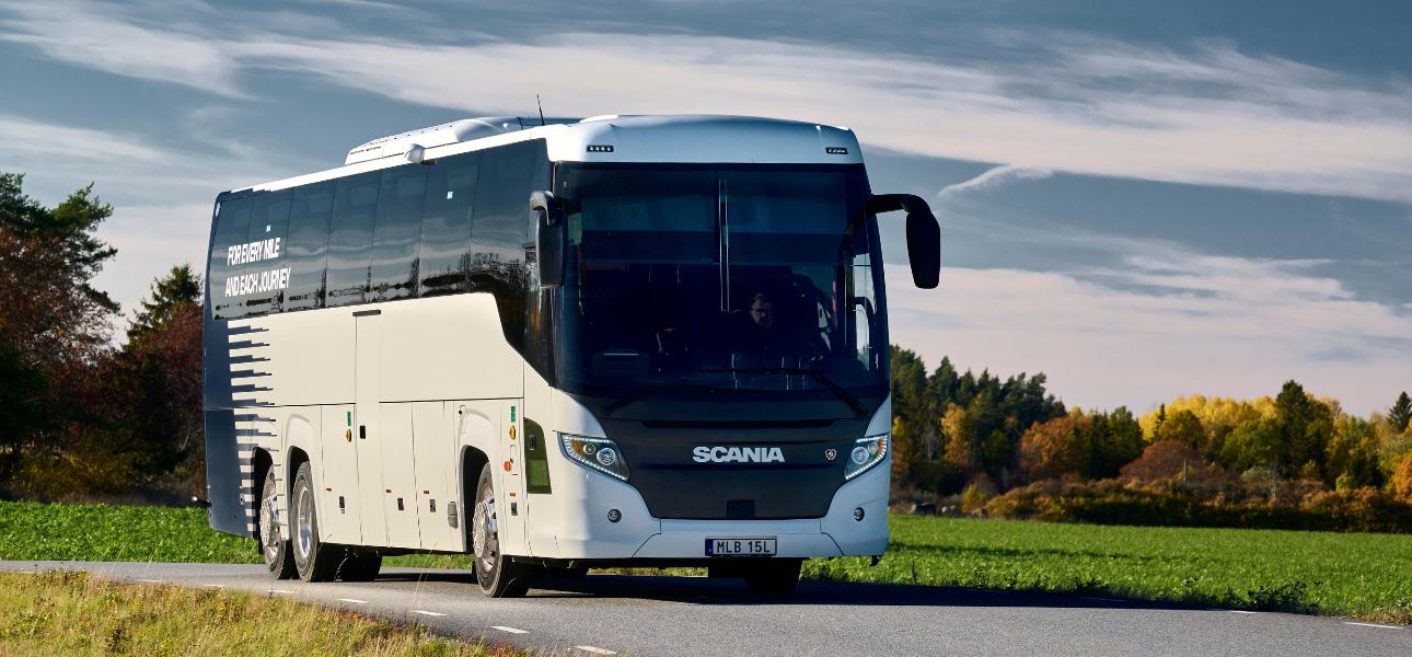 A Scania bus drives down a rural road, flanked by lush greenery and open fields.