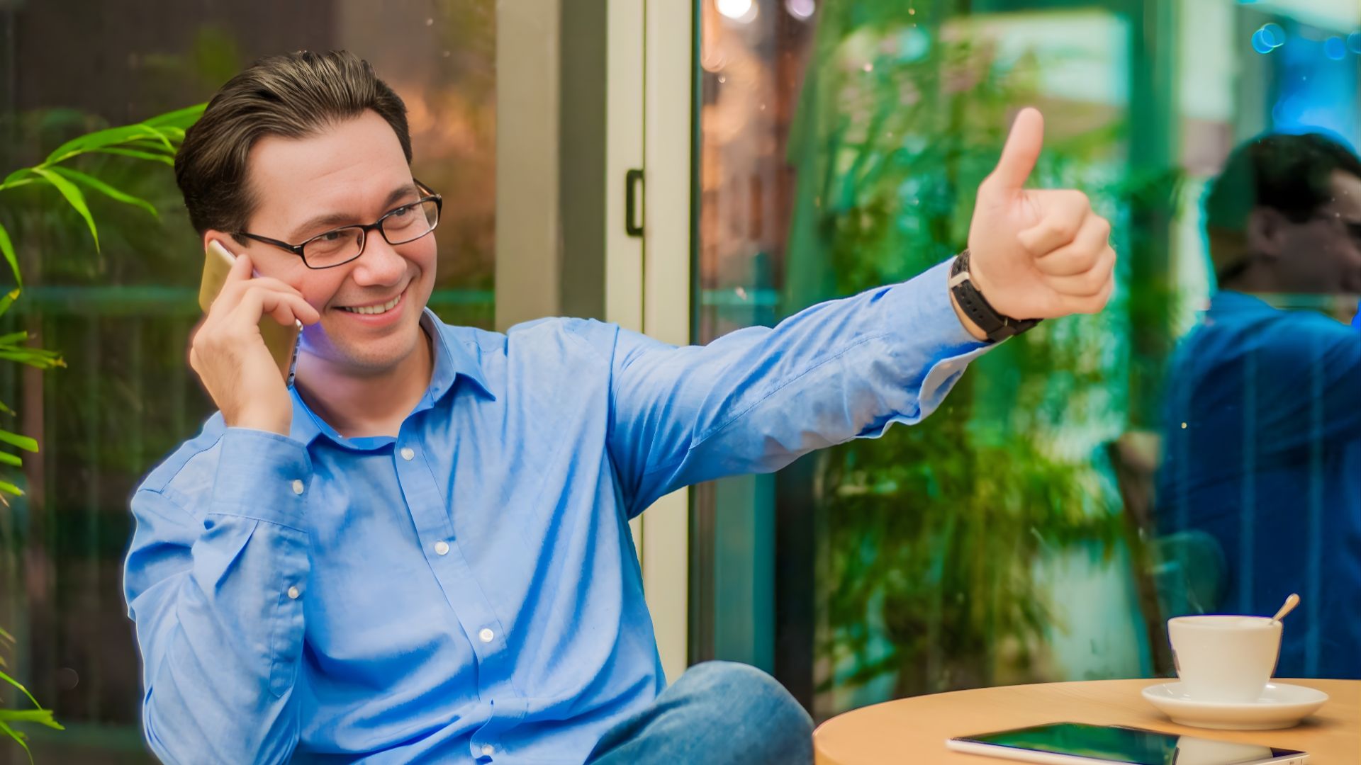 A man wearing glasses sits at a table, smiling and giving a thumbs up.