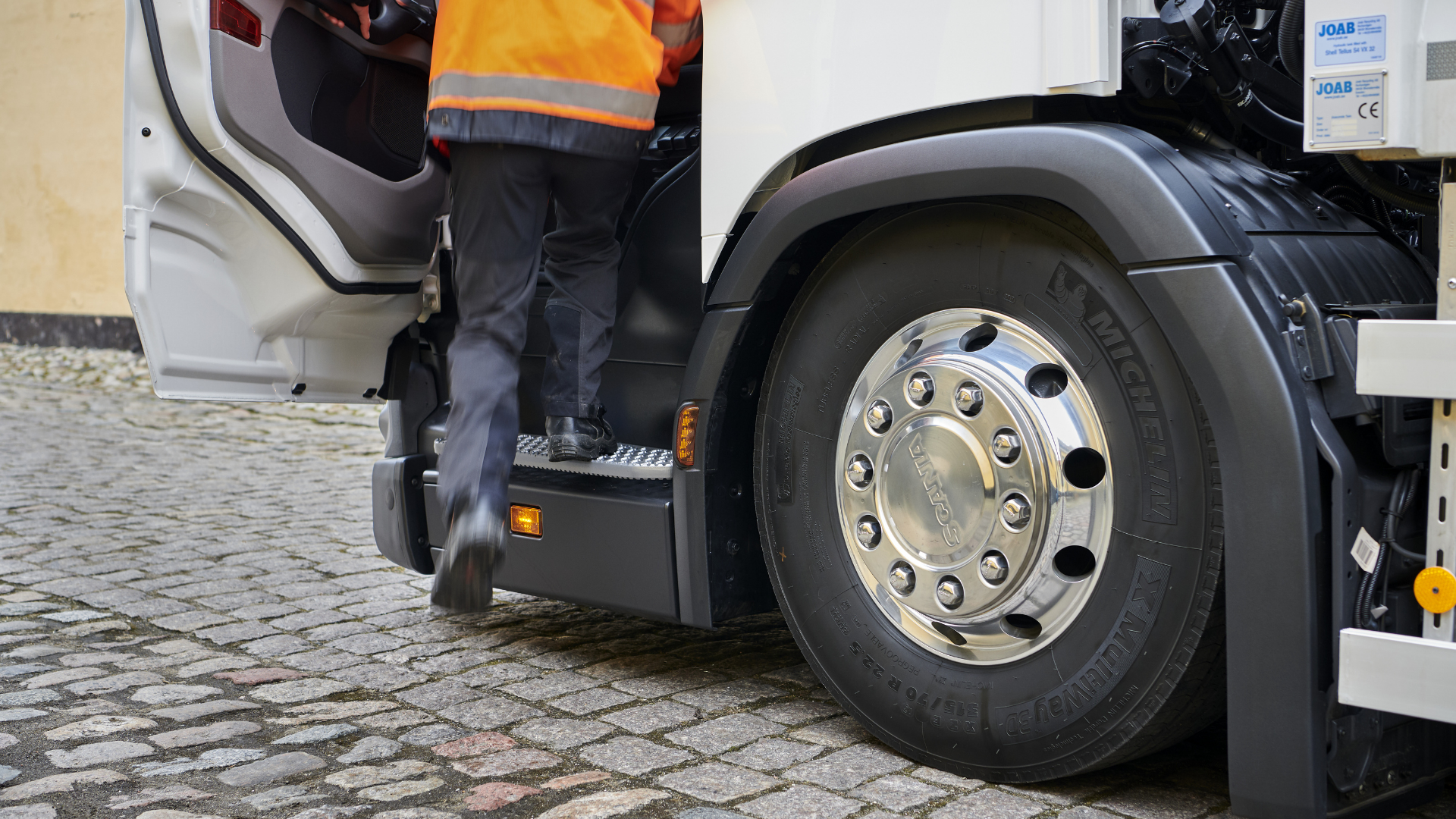 A filipino truck driver in an orange safety jacket steps into a white truck on a cobblestone street.