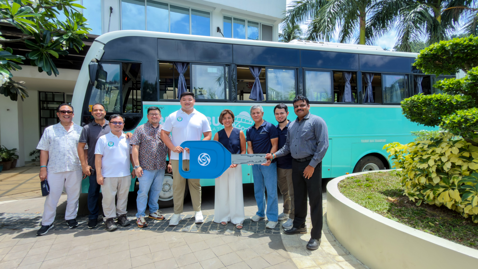 A group of ten people stand smiling in front of a large, turquoise bus.