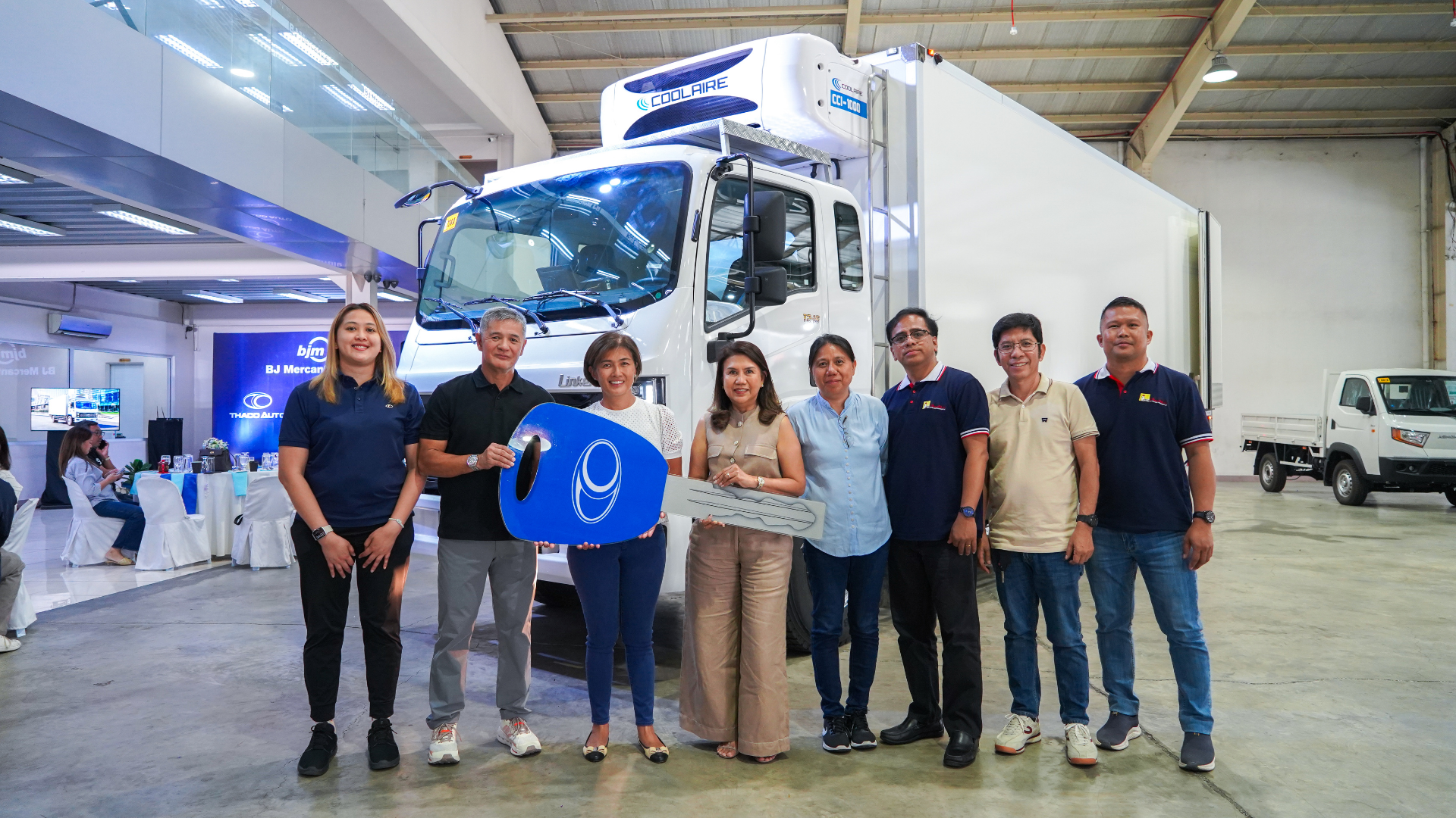 A group of eight people smile in front of a large white THACO Linker truck indoors.