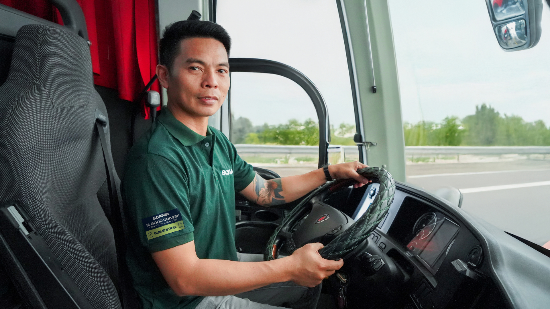A person in a green shirt confidently drives a bus on a sunny day, with a focused expression.