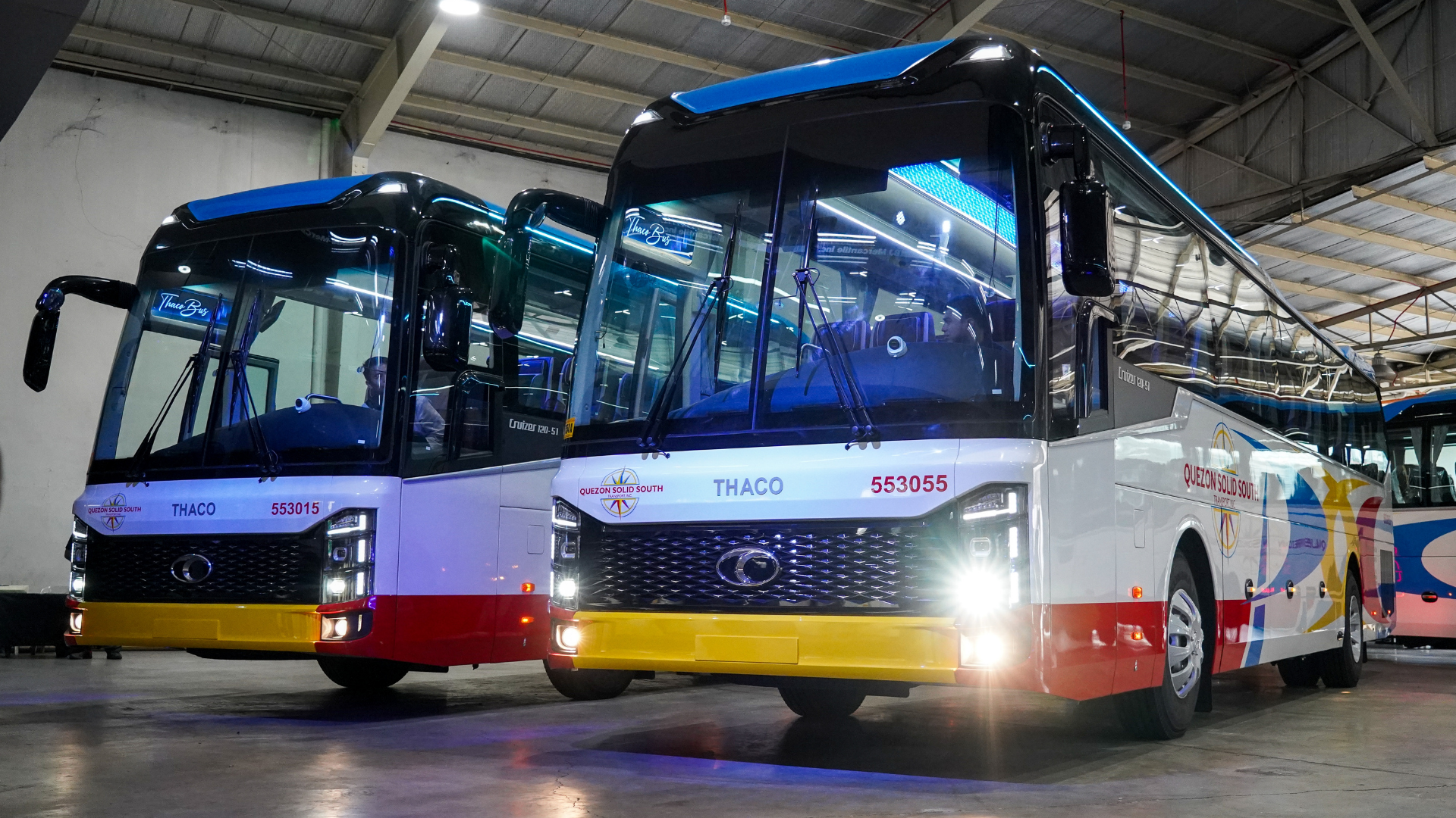 Front angle view of two brand new THACO passenger buses featuring the Quezon Solid South Transport livery, parked indoors with headlights illuminated.