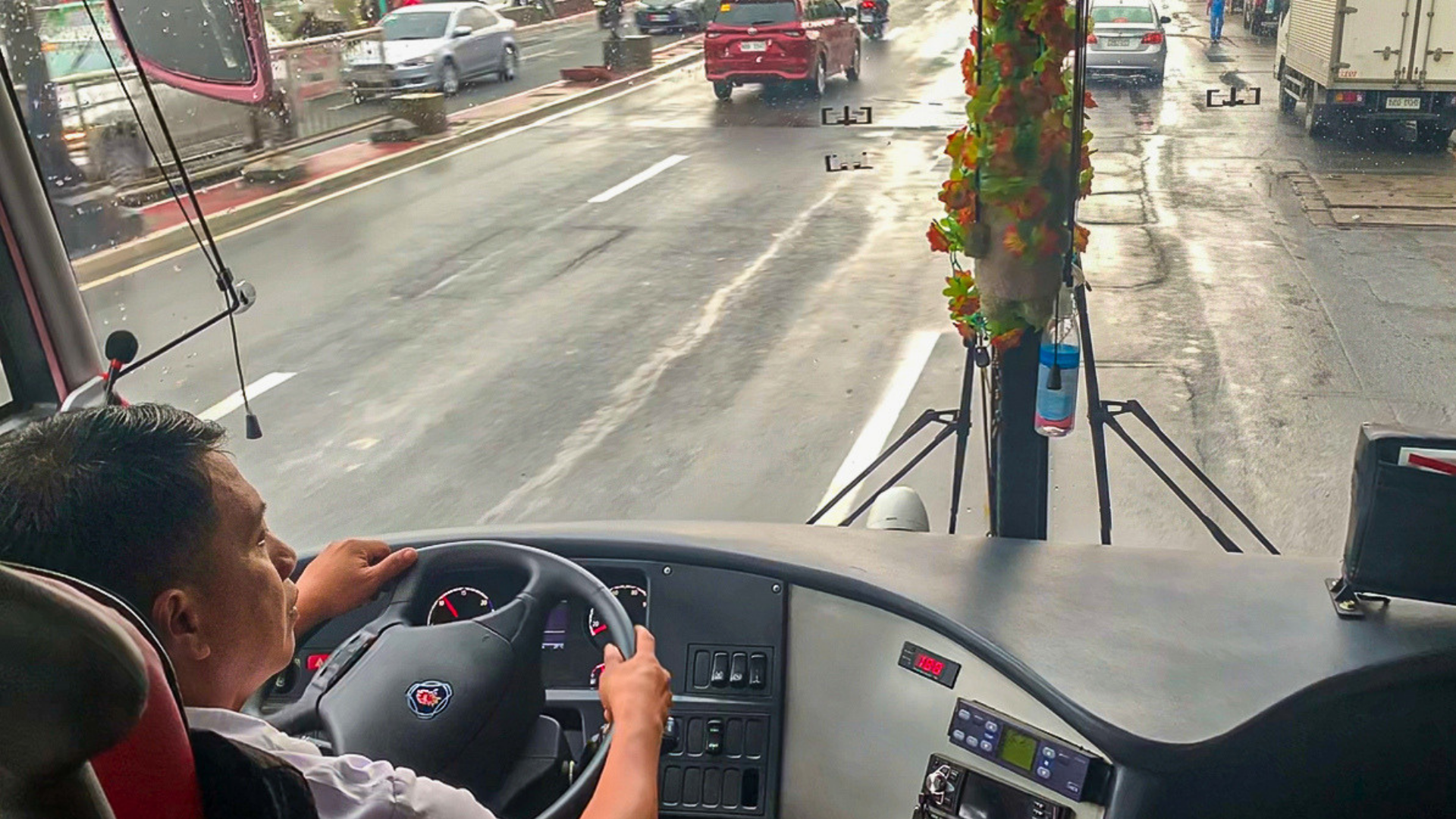 Interior view from behind a public transit bus driver navigating through urban city traffic on a wet street.