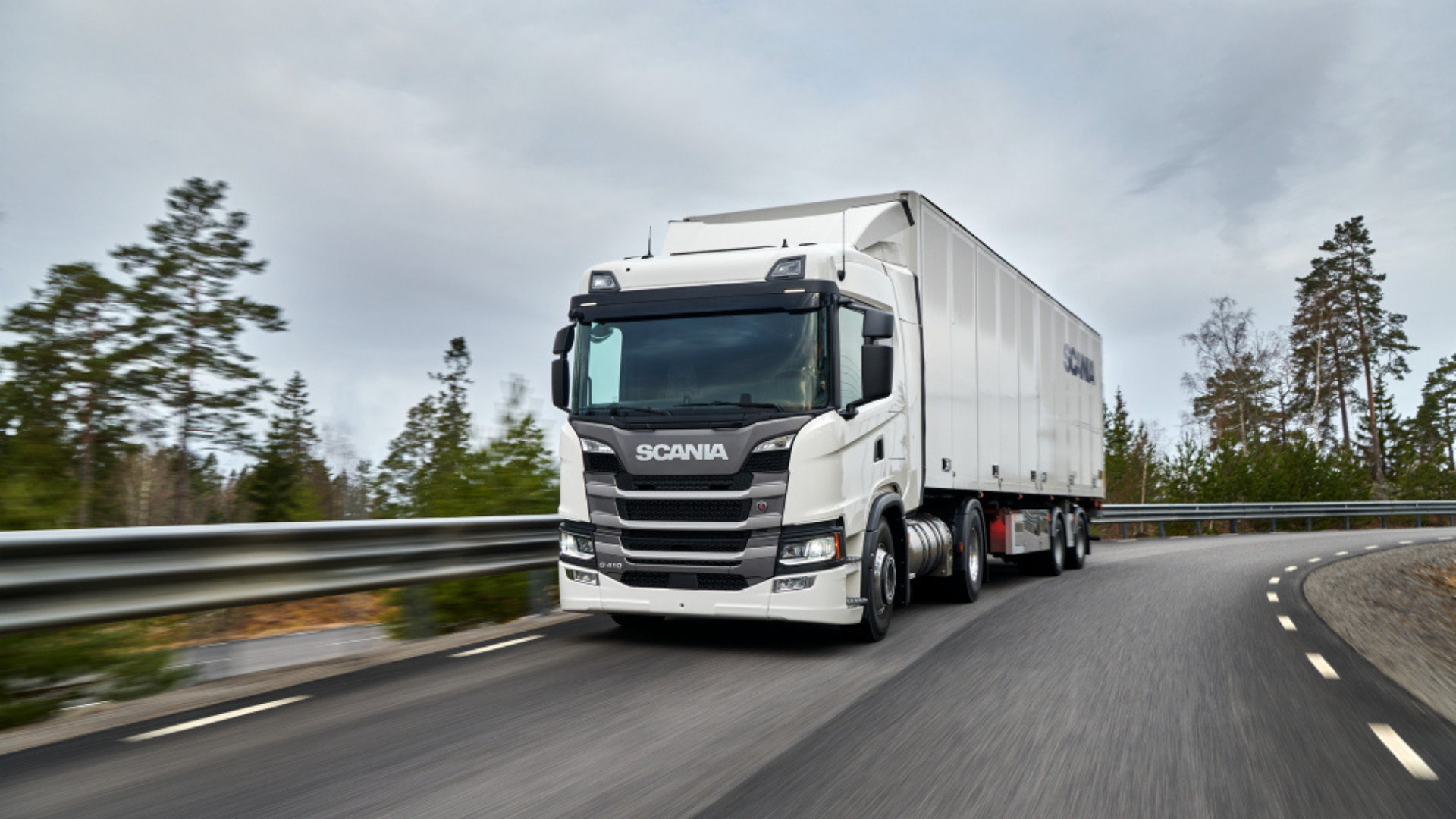 A white Scania commercial semi-truck driving along a curved paved highway bordered by trees under a cloudy sky.